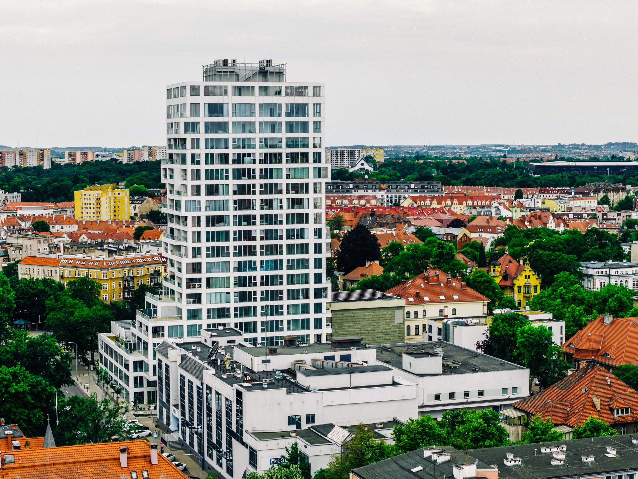 Biały wieżowiec wśród miejskiej zabudowy widziany z powietrza.