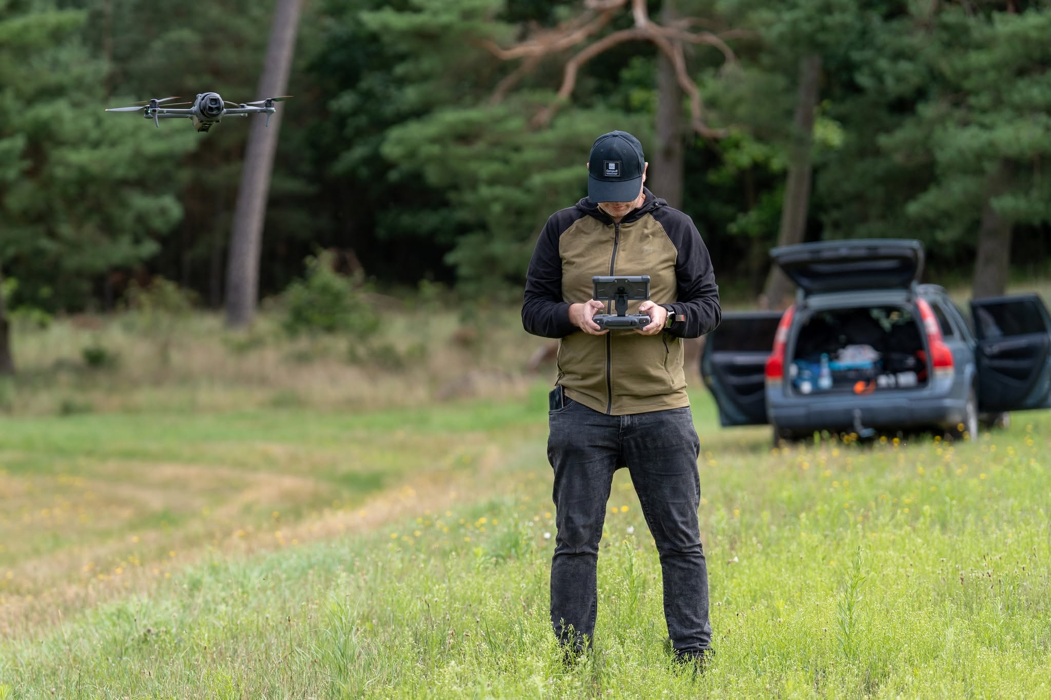 Operator steruje dronem na łące, w tle otwarty samochód i drzewa.