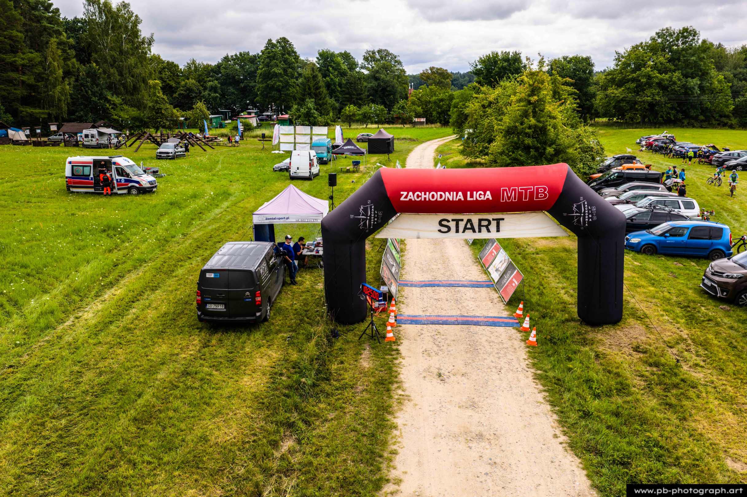 Ujęcie z drona na start wyścigu MTB w Szczecinie, widok na bramę startową oraz otoczenie z zielenią i pojazdami.