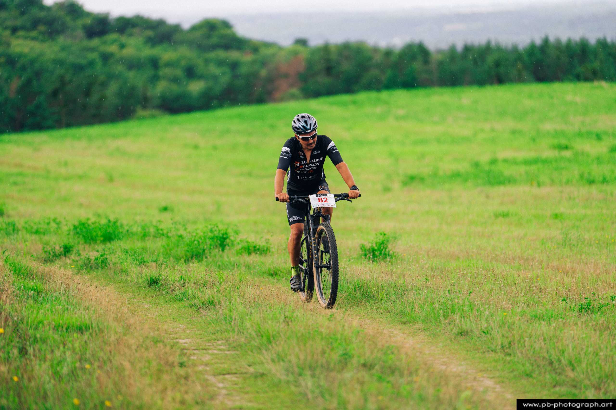 Ujęcie z drona zawodnika MTB na trasie wyścigu w Szczecinie, otoczony zielonymi łąkami i drzewami.