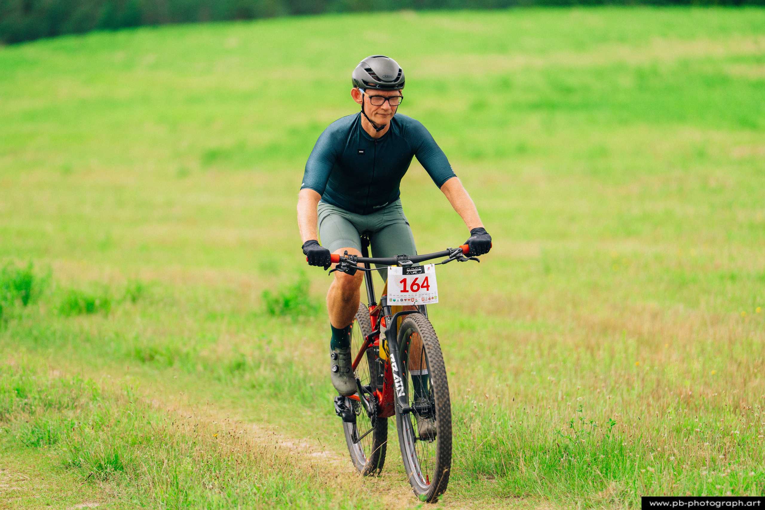 Zawodnik na rowerze górskim podczas wyścigu MTB w Szczecinie, z numerem startowym 164 na tle zielonych pól.