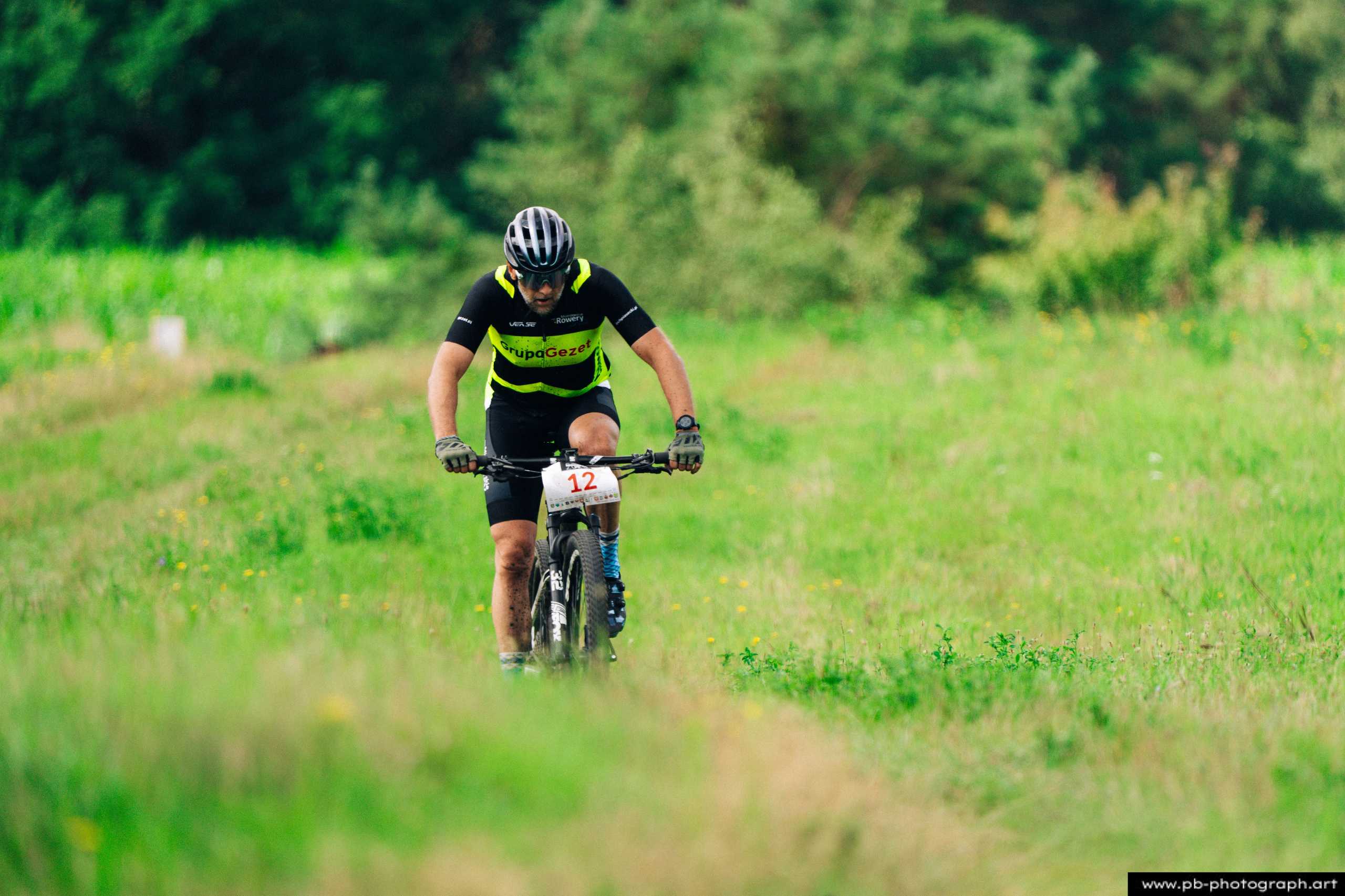 Fotografia z drona przedstawiająca zawodnika MTB na trasie wyścigu w Szczecinie, zmierzającego wzdłuż zielonej ścieżki.