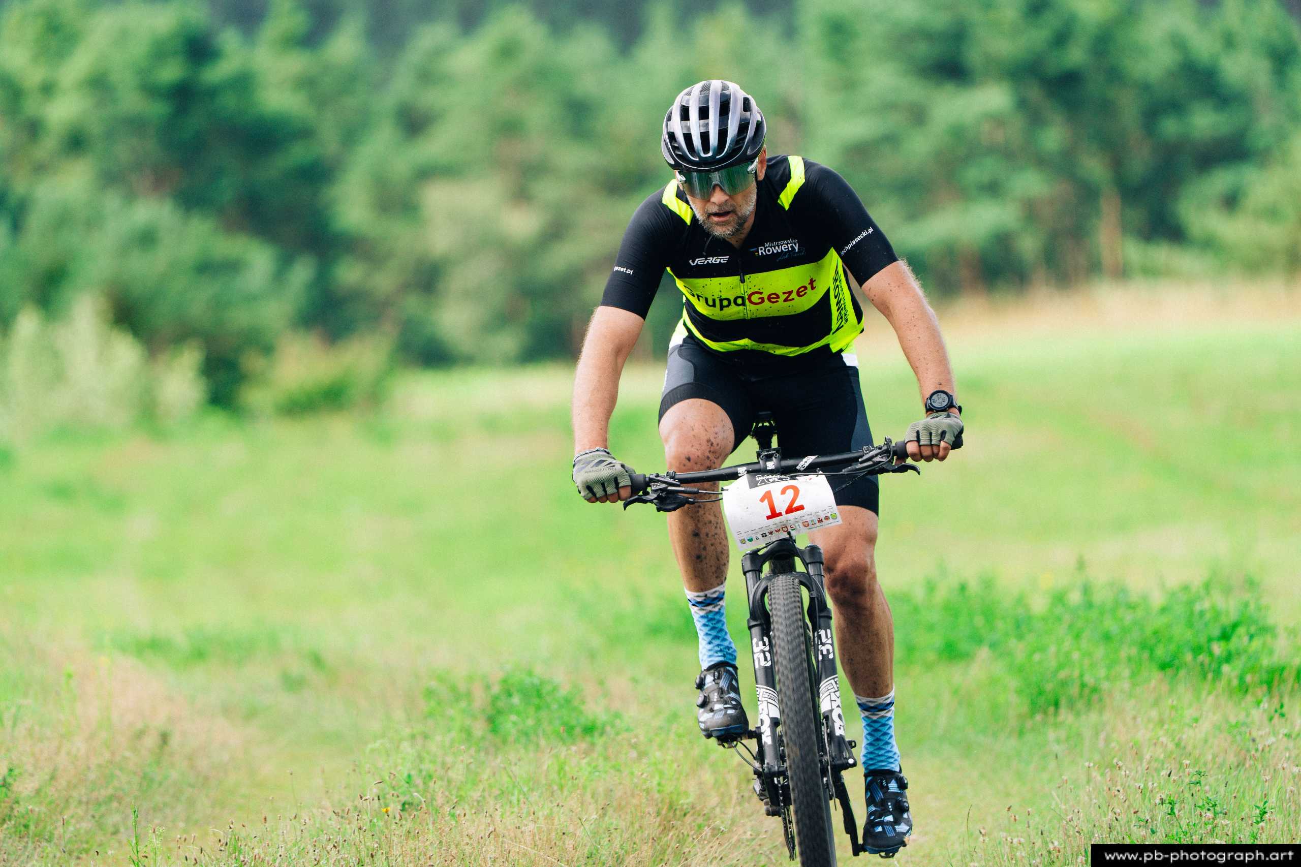 Zawodnik na rowerze górskim z numerem 12 podczas wyścigu MTB w Szczecinie, w tle zieleń lasu.