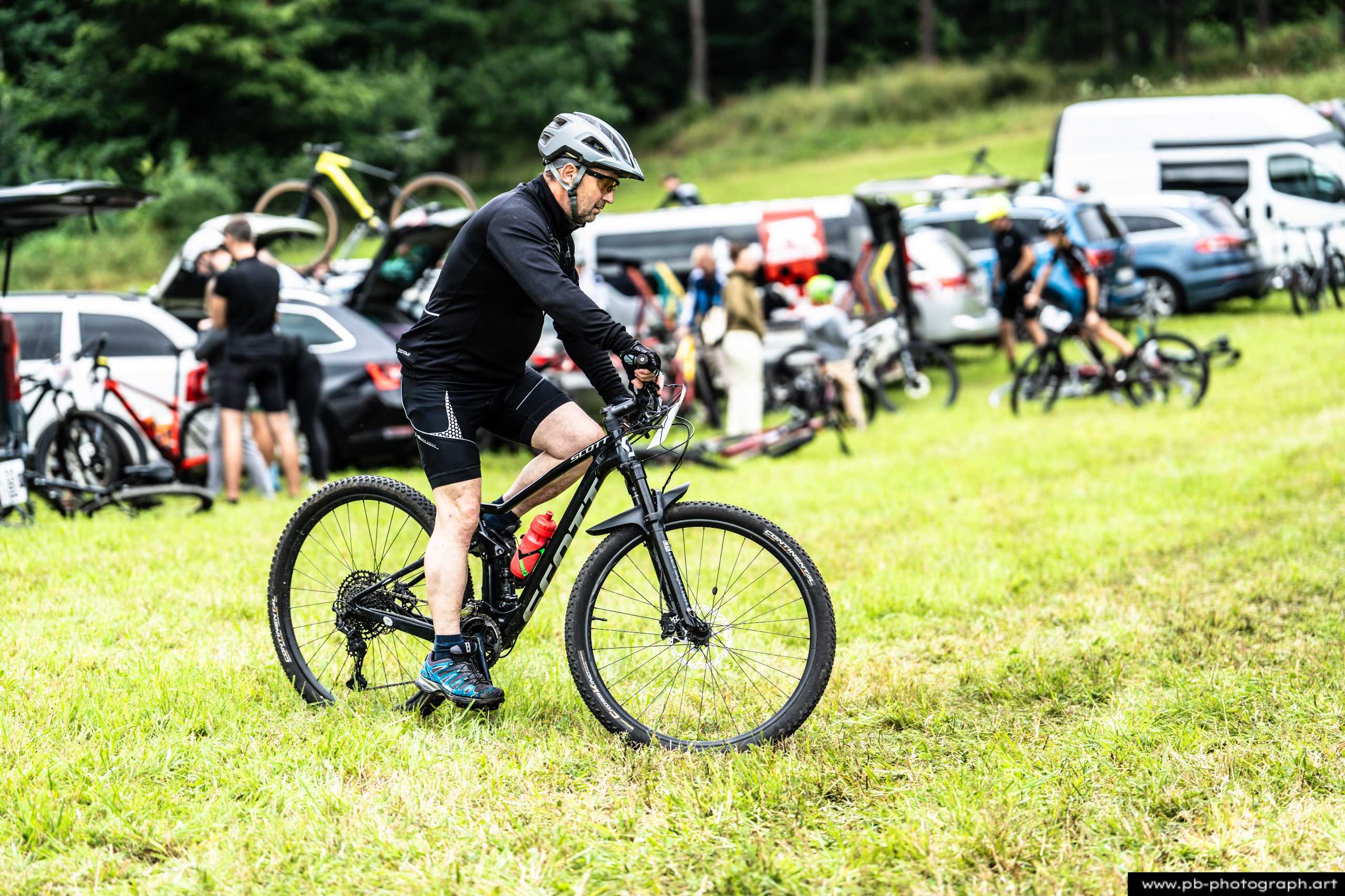 Zawodnik na rowerze górskim przygotowujący się do startu w wyścigu MTB w Szczecinie, z tłem samochodów i zawodników.