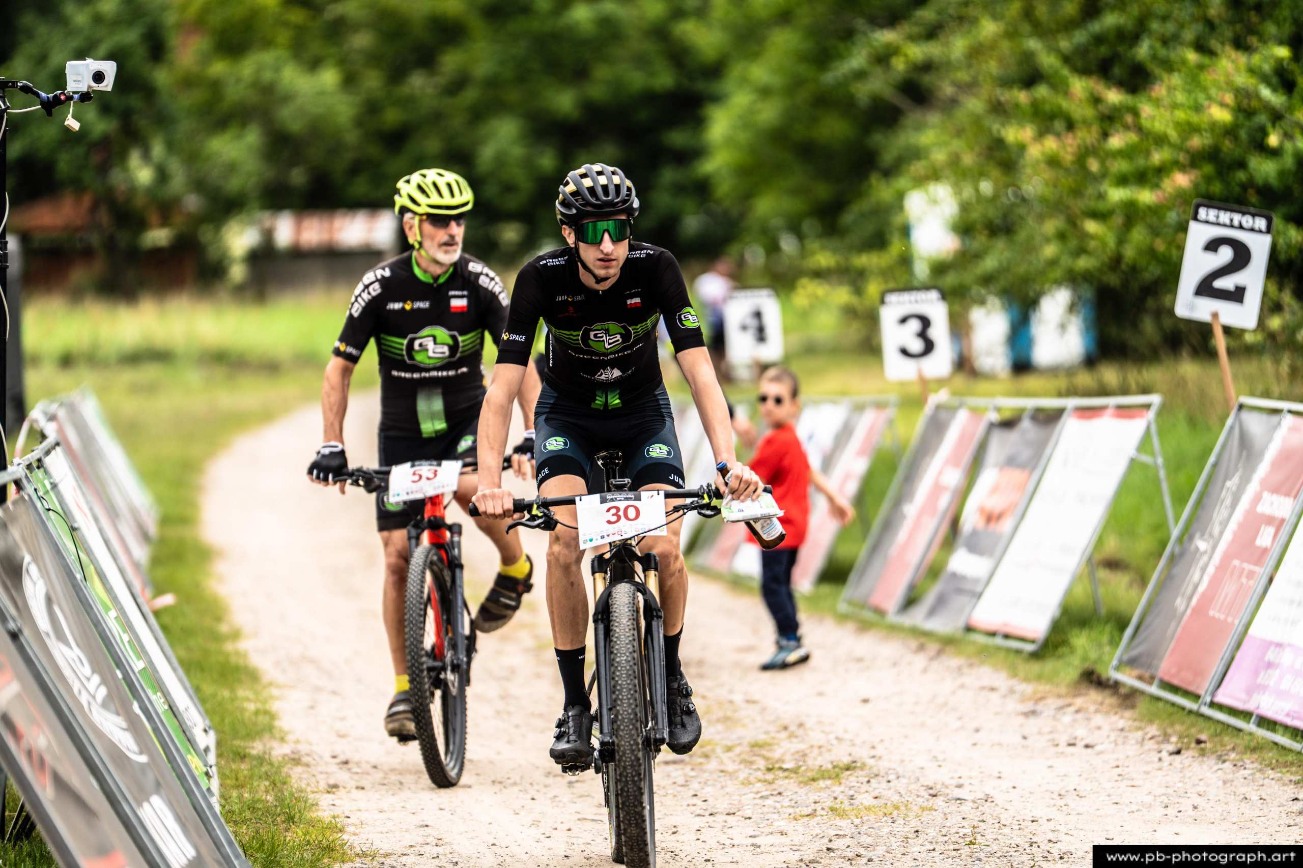 Zawodnicy MTB na trasie wyścigu w Szczecinie, z numerami startowymi i dzieckiem w tle.