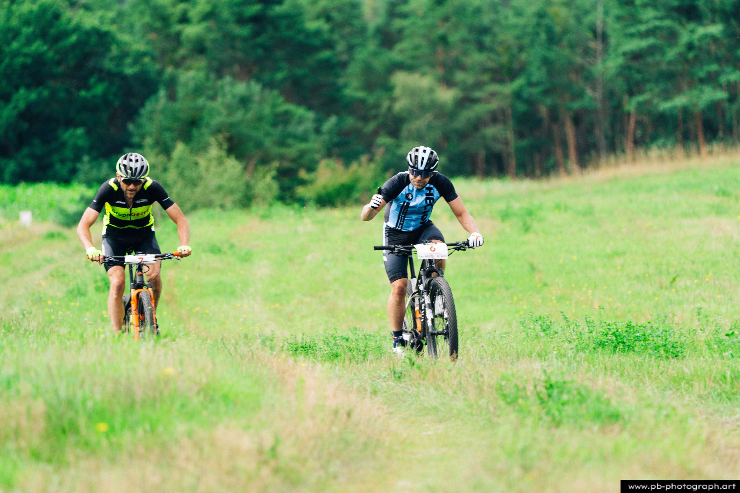Ujęcie z drona dwóch zawodników MTB na trasie wyścigu w Szczecinie, otoczeni zielonymi łąkami i lasem.