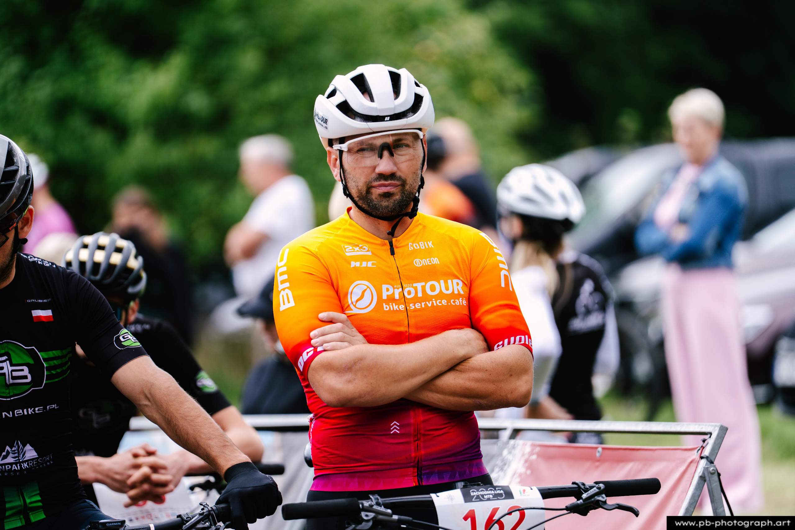 Zawodnik w pomarańczowej koszulce na tle innych uczestników wyścigu MTB w Szczecinie, czekający na start.