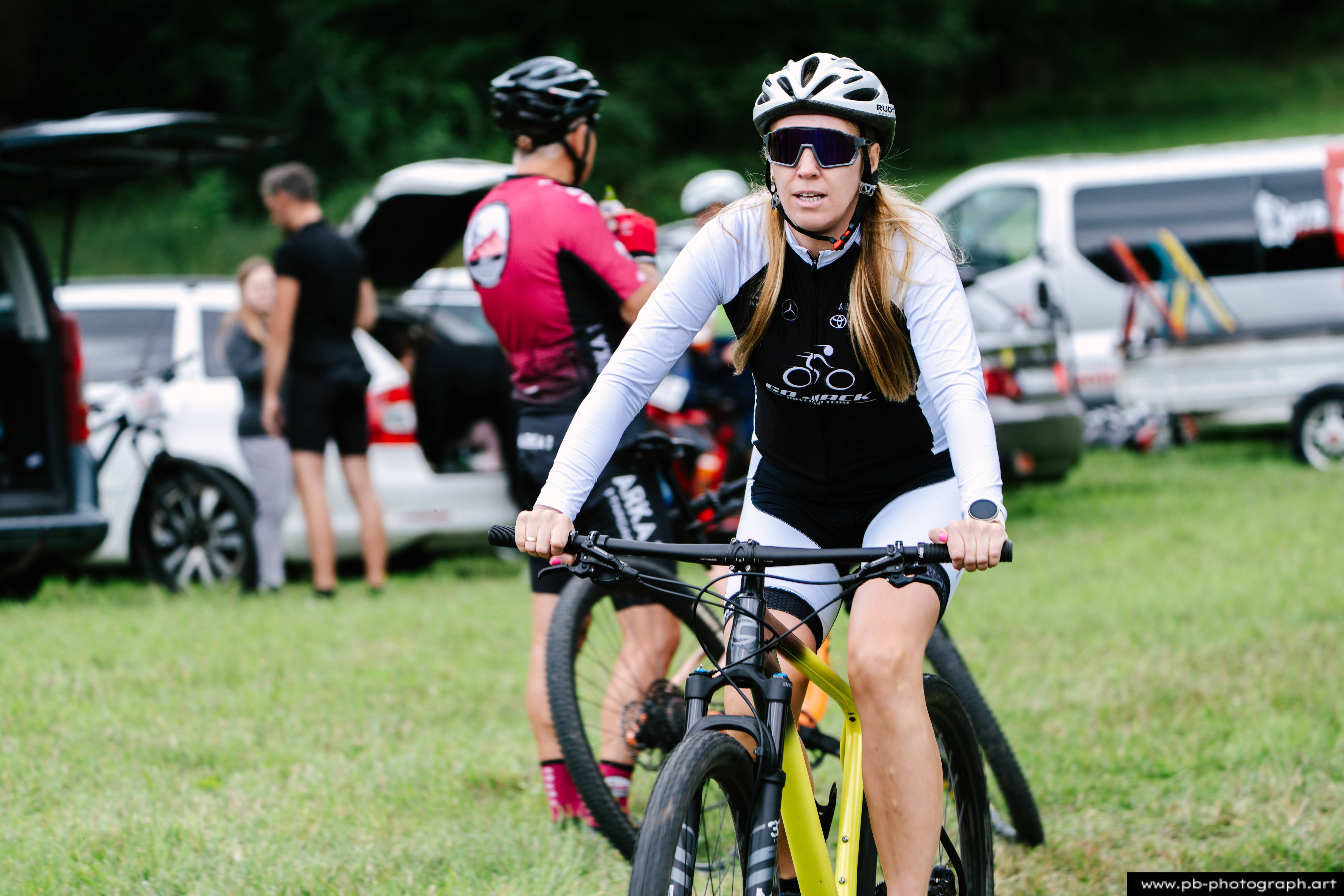 Zawodniczka na rowerze górskim przygotowująca się do wyścigu MTB w Szczecinie, z innymi uczestnikami w tle.