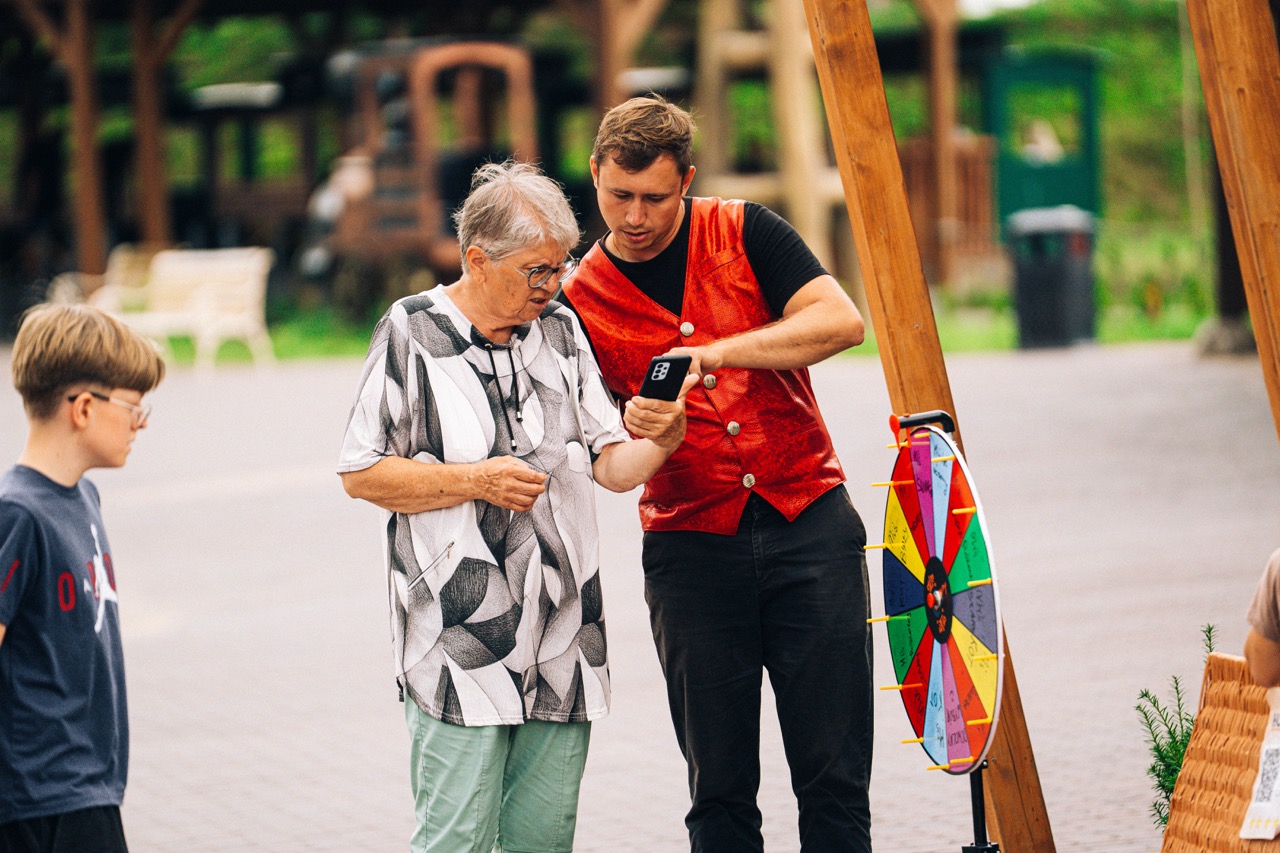 Iluzjonista w czerwonej kamizelce pokazuje starszej kobiecie telefon, obok stoi chłopiec i kolorowe koło fortuny.