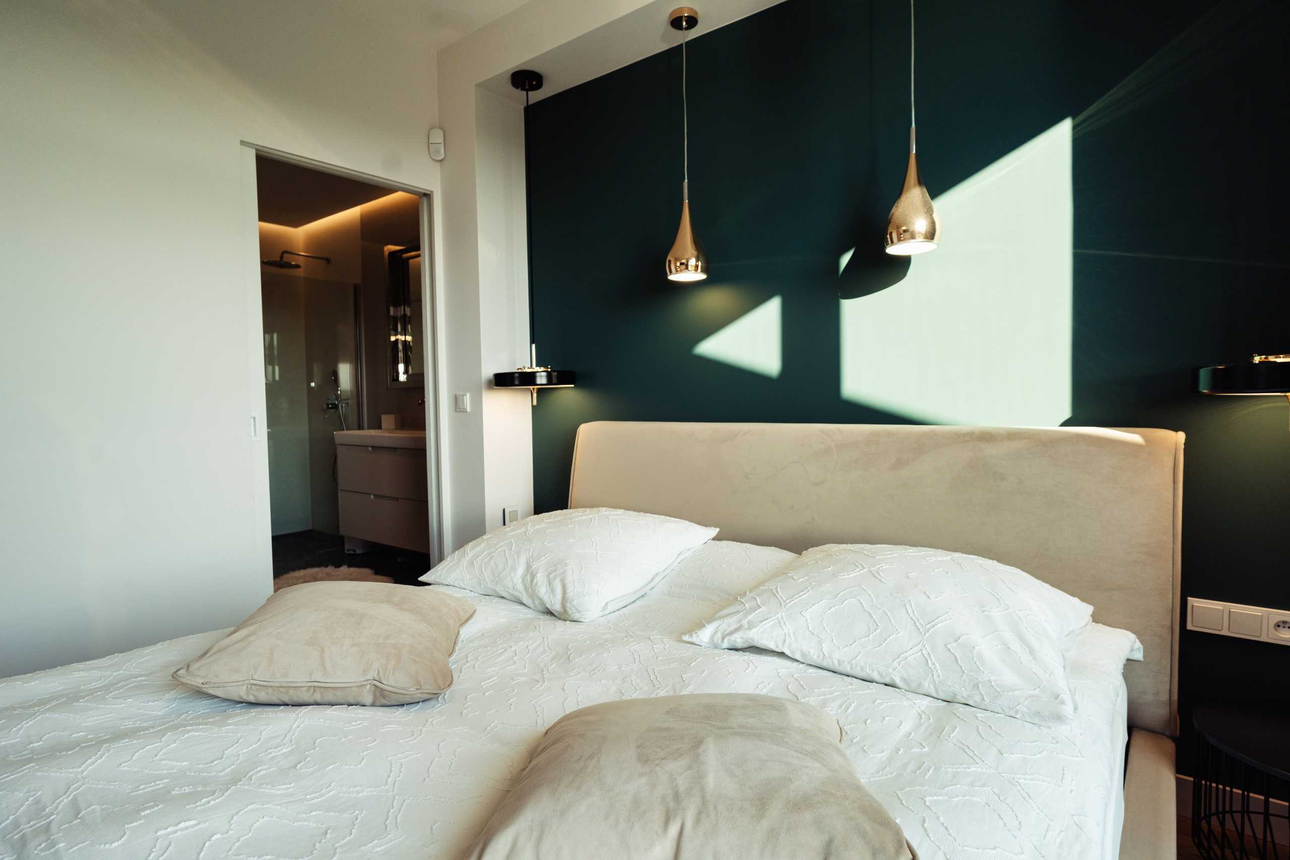 Sypialnia w apartamencie w Warszawie z eleganckim łóżkiem i nowoczesnymi lampami na tle zielonej ściany.
