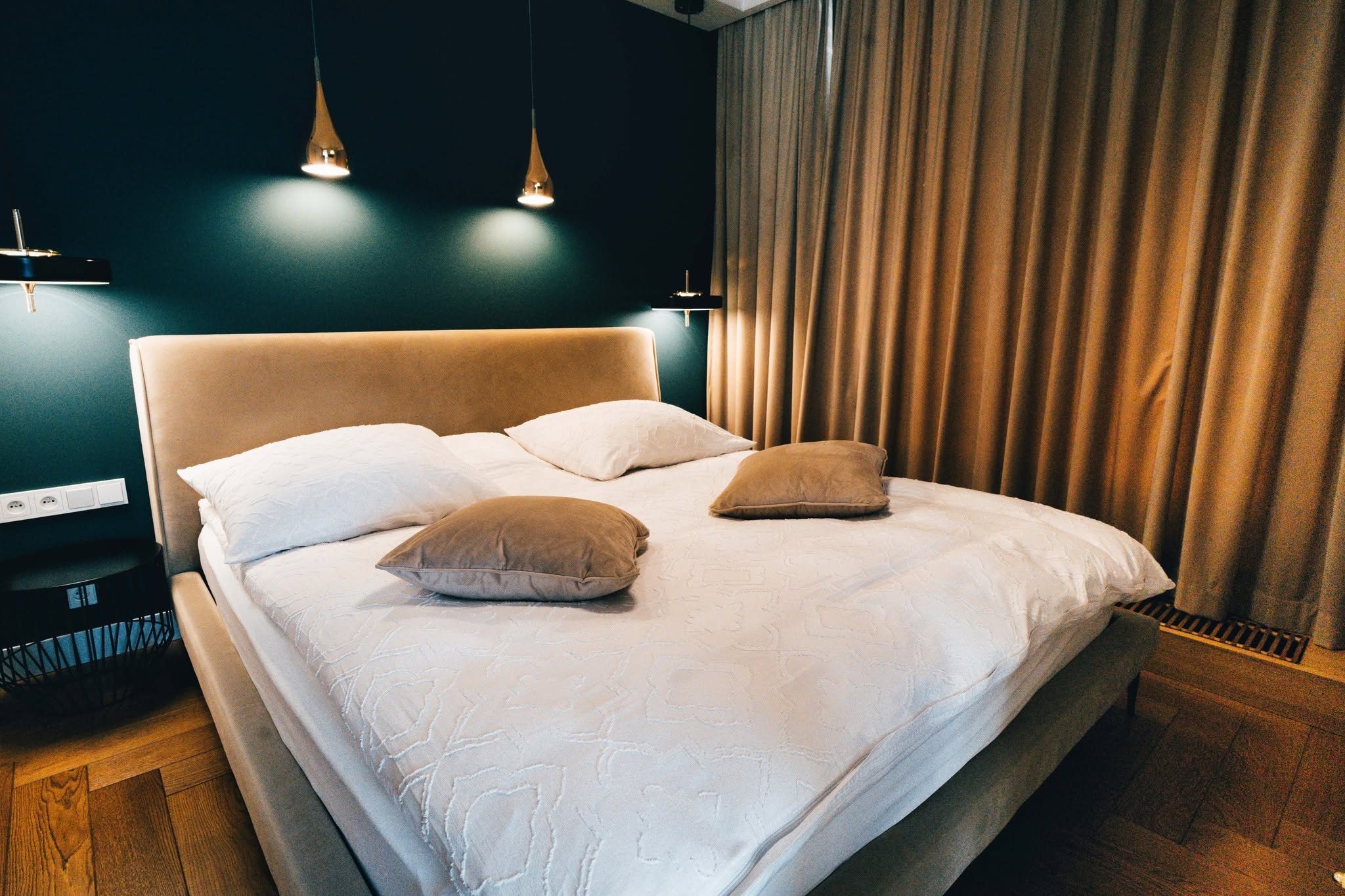 Eleganckie łóżko w apartamencie z poduszkami na tle ciemnej ściany i zasłon z naturalnych materiałów.