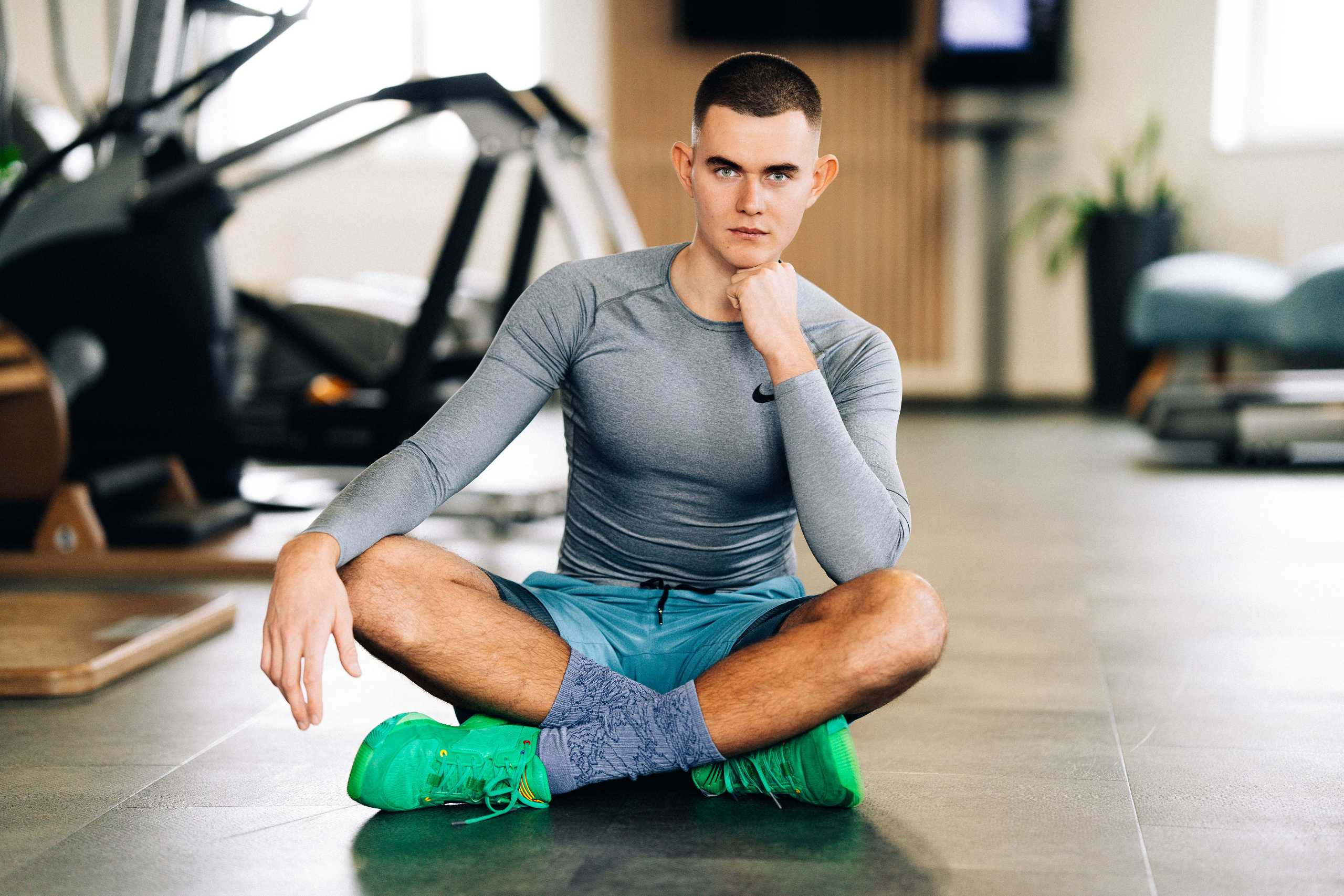 Młody mężczyzna w sportowym stroju siedzi na podłodze w klubie fitness, prezentując pewność siebie i zaangażowanie.