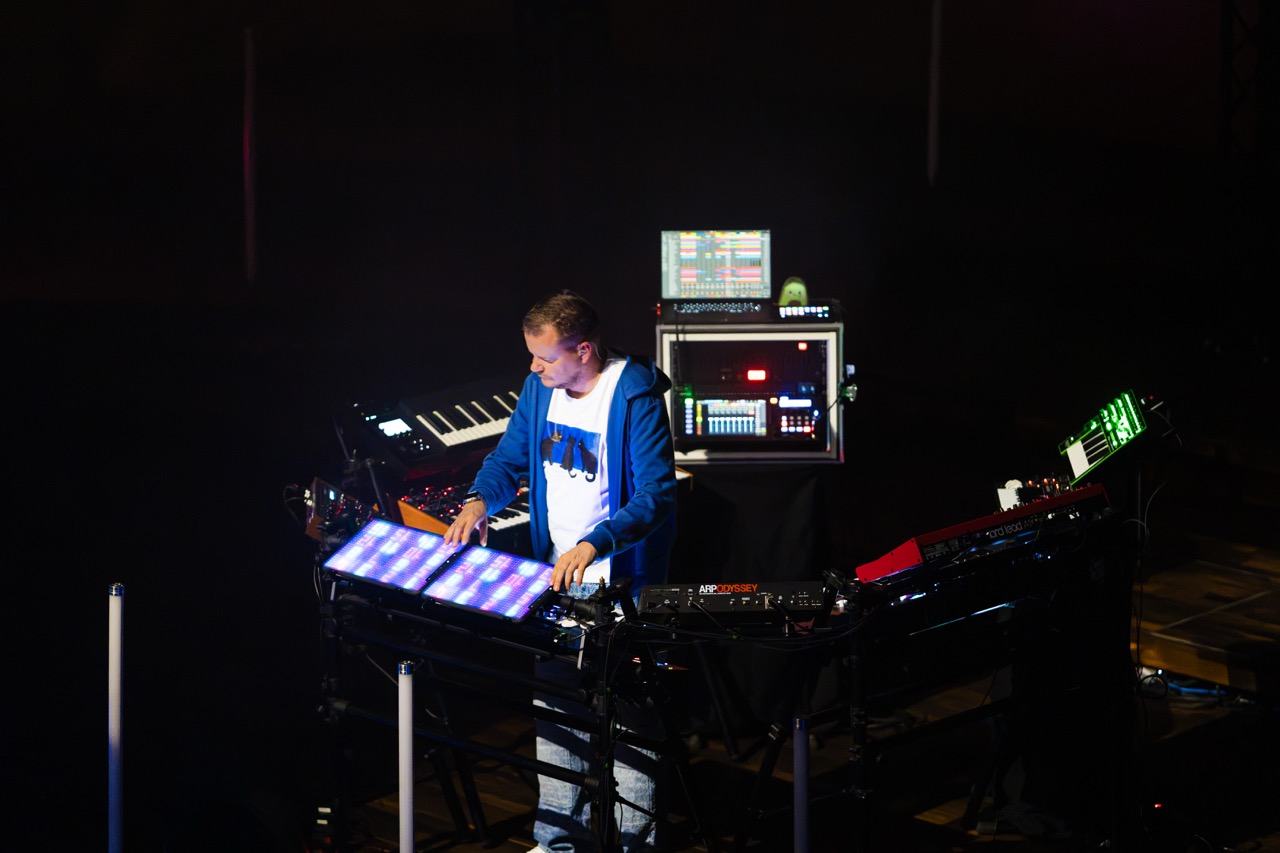 Muzyk Madis podczas występu w Filharmonii Szczecińskiej, z instrumentami elektronicznymi w ciemnej scenerii.