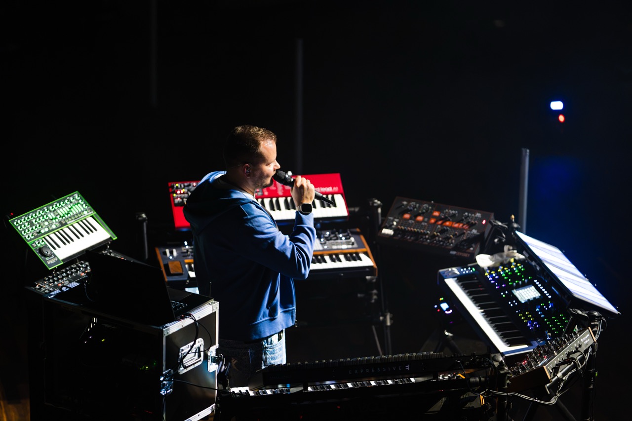 Muzyk z mikrofonem w ręku, otoczony instrumentami elektronicznymi podczas koncertu w Filharmonii Szczecińskiej.