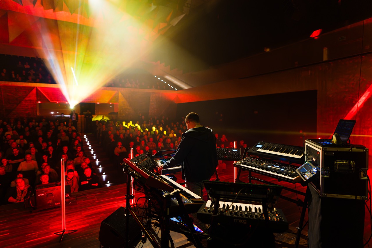 Artysta Madis na scenie w Filharmonii Szczecińskiej, z widownią w tle oraz instrumentami elektronicznymi.