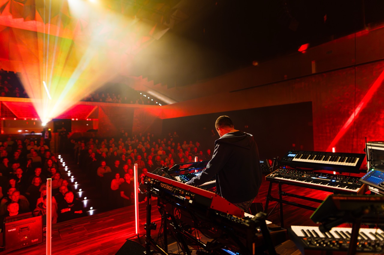 Fotografia koncertowa w Filharmonii Szczecińskiej — kompozytor Madis przy instrumentach, w tle oświetlona publiczność.