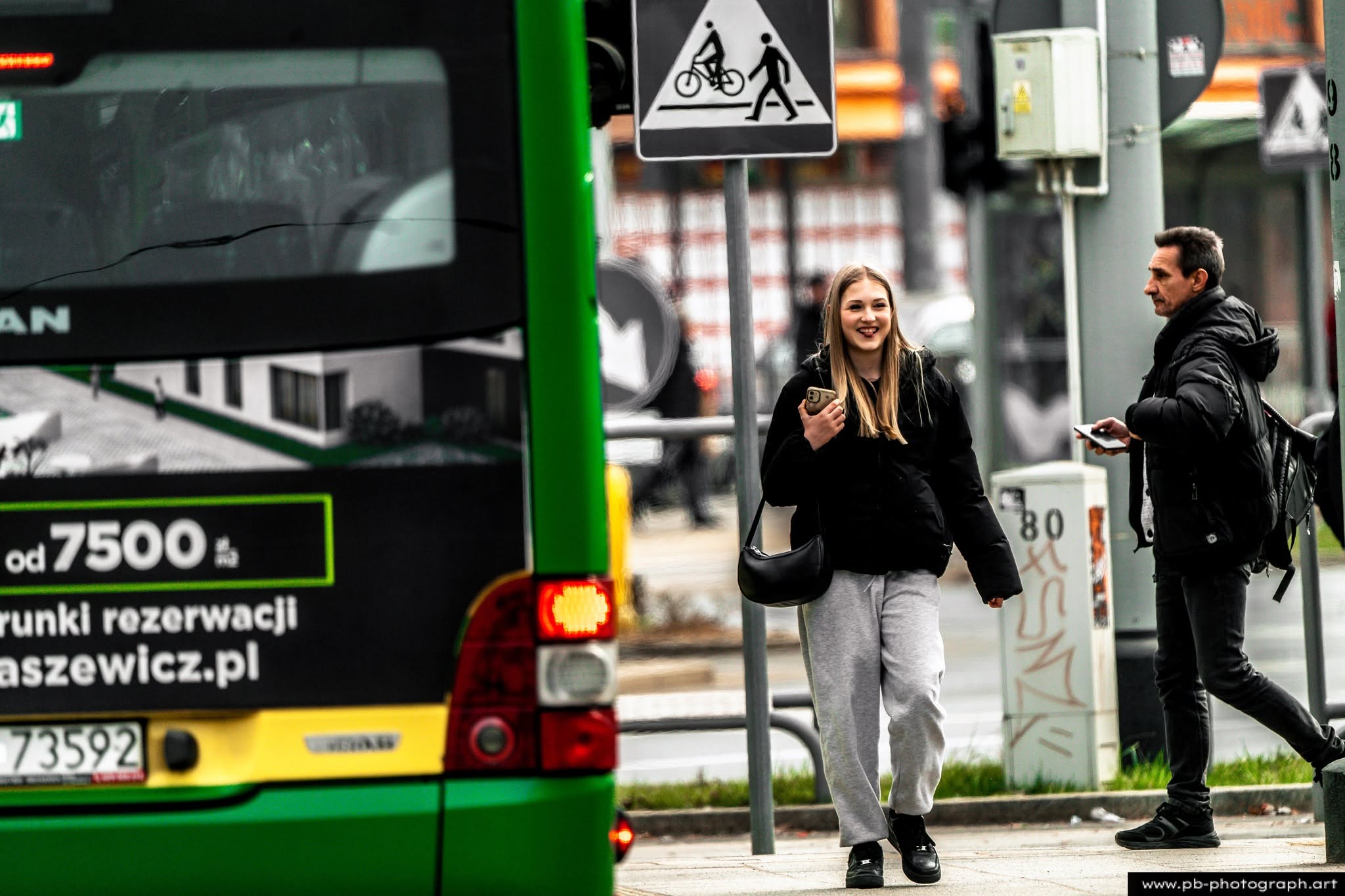 Ujęcie z ulicy w Szczecinie — przechodnie obok autobusu, kobieta z telefonem w ręku.