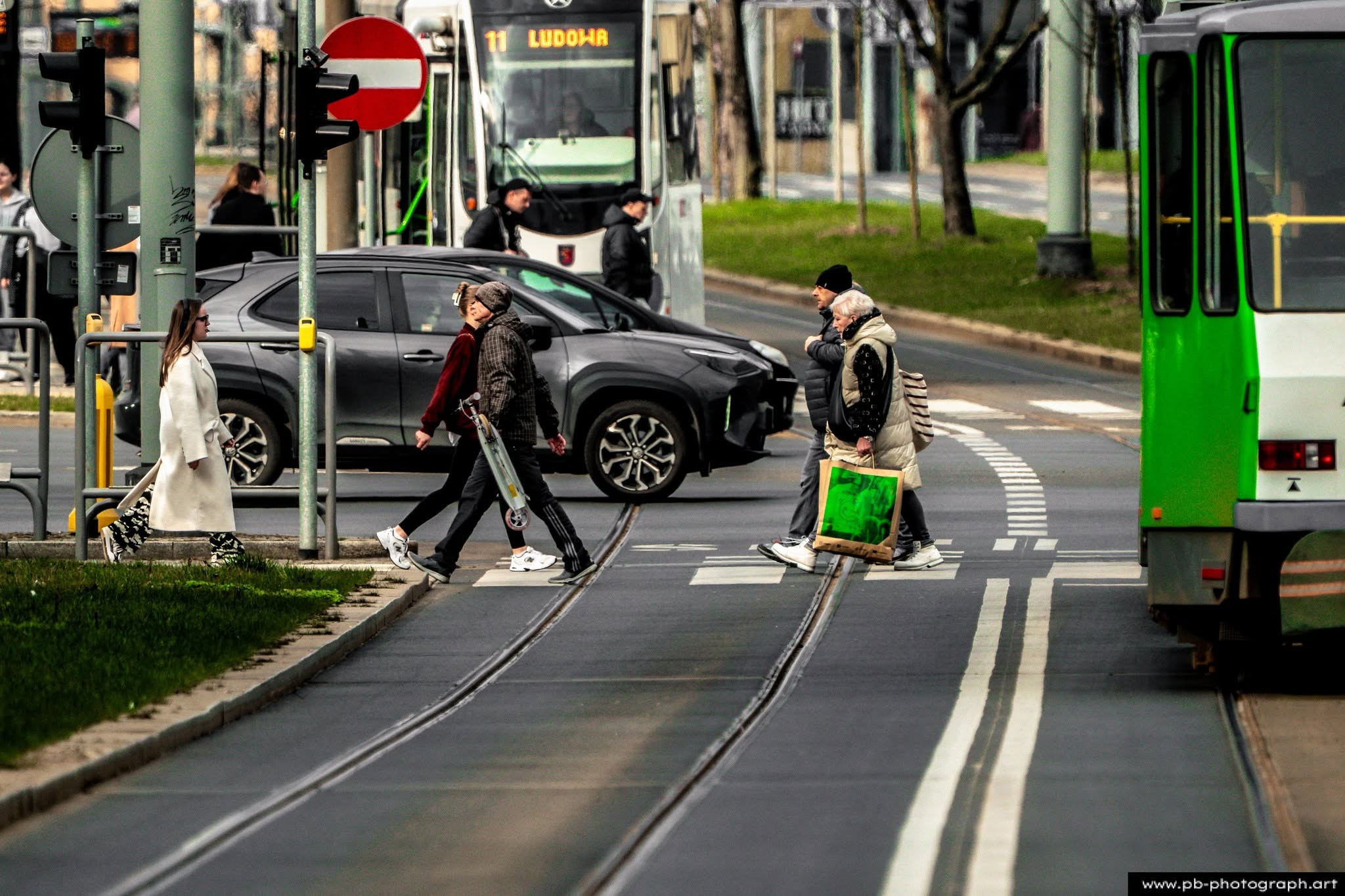 Przechodnie na pasach w Szczecinie, obok tramwajów i samochodów, uchwycone z perspektywy ulicznej.