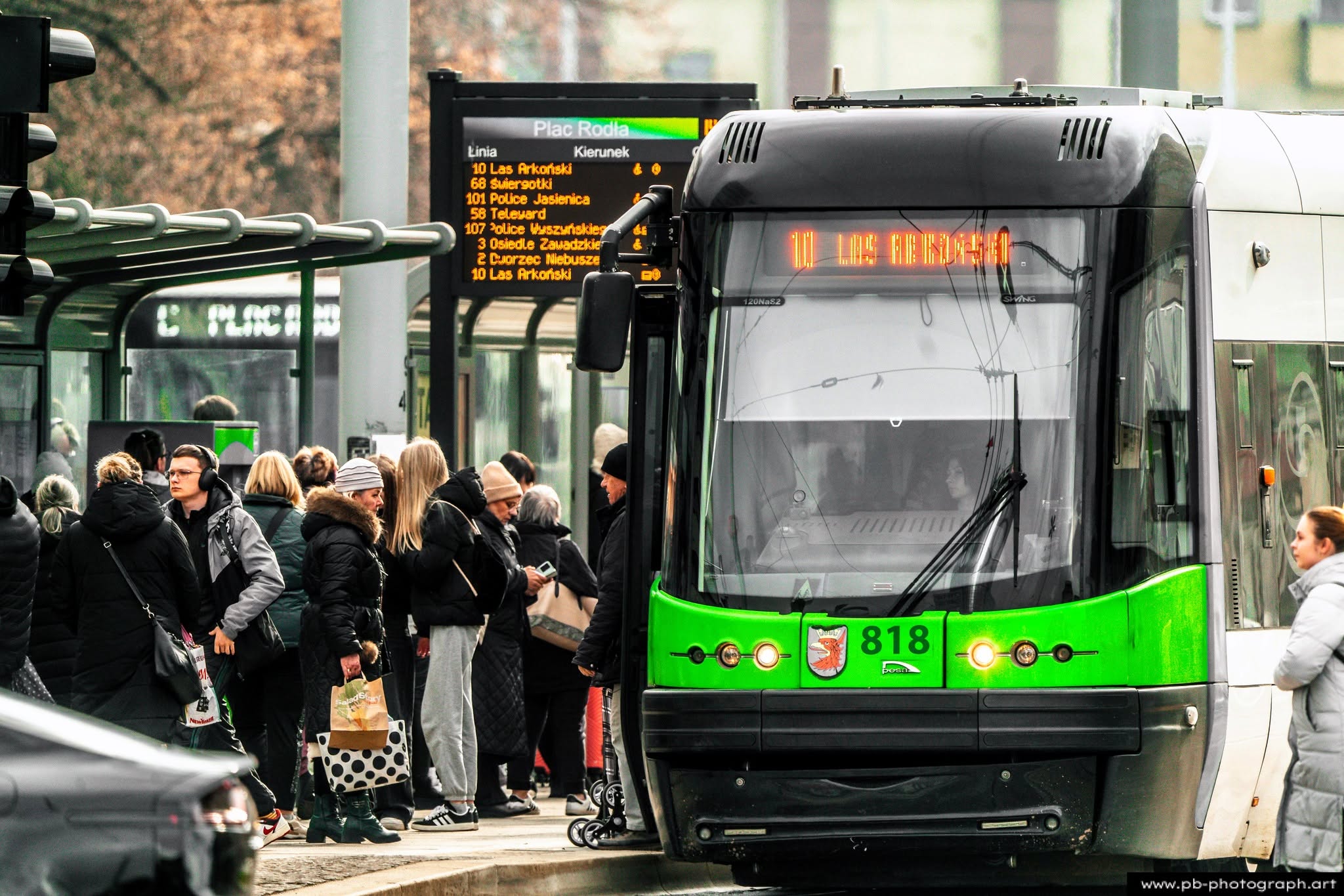 Tramwaj w Szczecinie zatrzymany na przystanku, pasażerowie oczekują na przyjazd, w tle tablica z rozkładem jazdy.