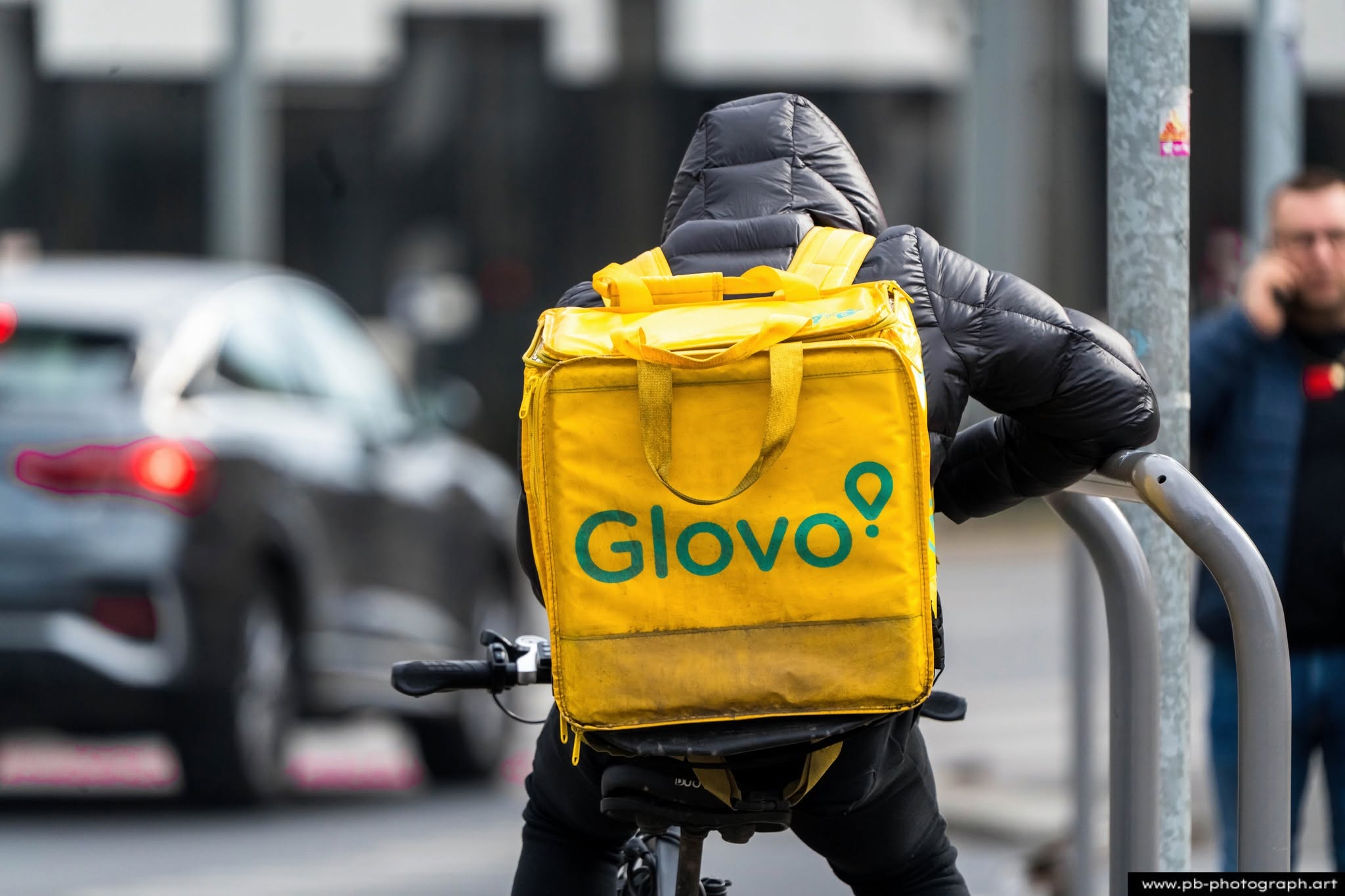 Kurier na rowerze z torbą Glovo w Szczecinie, czekający na zmianę świateł na ruchliwej ulicy.