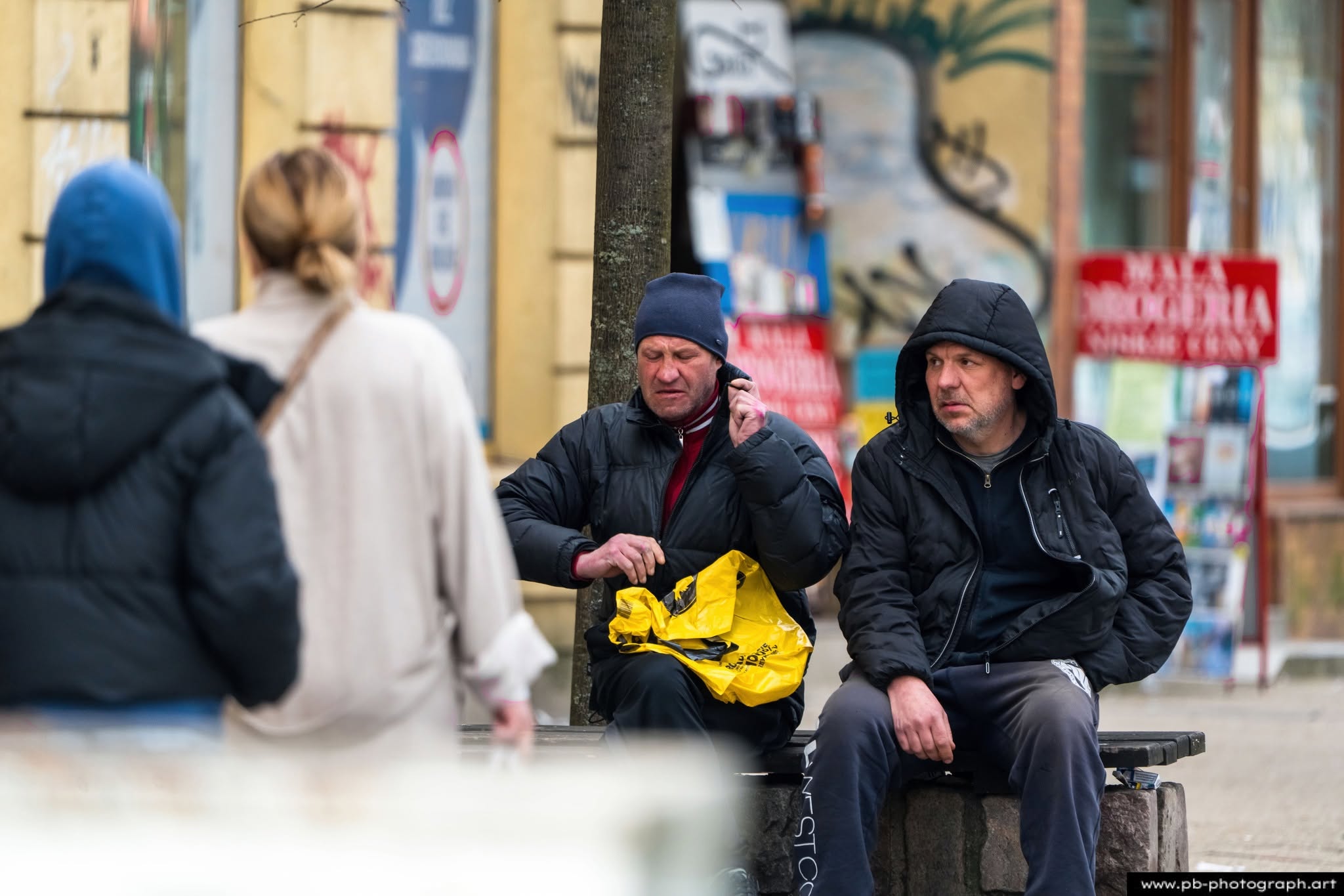 Mężczyźni w odzieży wierzchniej siedzący na ławce w Szczecinie, obserwujący przechodniów na ulicy.
