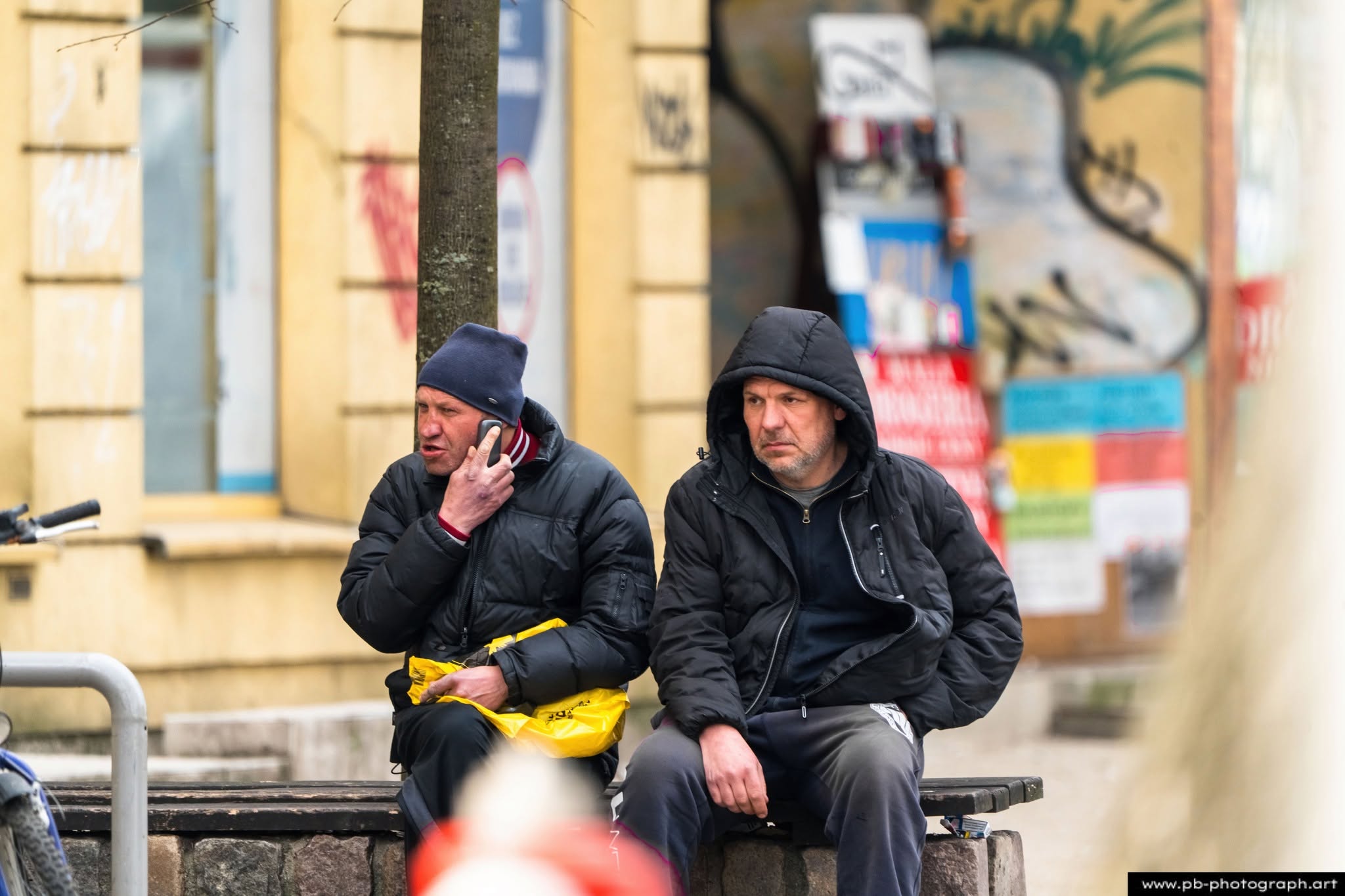 Dwaj mężczyźni siedzący na ławce w Szczecinie, jeden rozmawia przez telefon, drugi obserwuje otoczenie.