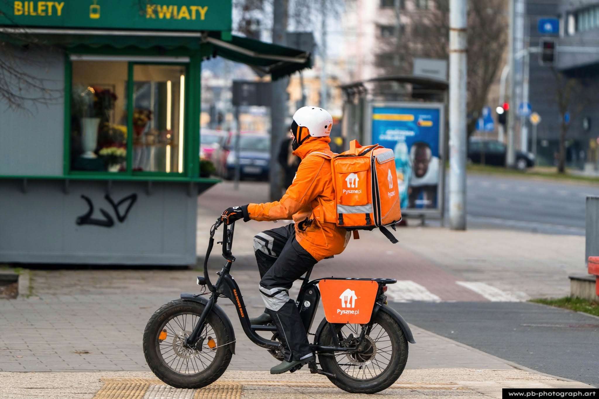 Kurier na rowerze elektrycznym w pomarańczowej kurtce, przejeżdżający obok kiosku w Szczecinie.