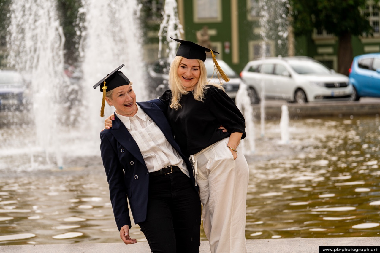 Reportaż z absolutorium w Szczecinie — dwie absolwentki w togach przy fontannie w Parku Kasprowicza.