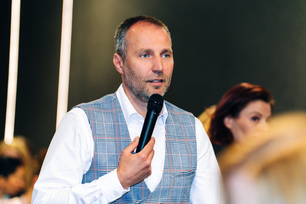 Mężczyzna w eleganckiej kamizelce przemawia z mikrofonem w piwnicy hotelu Fokus.