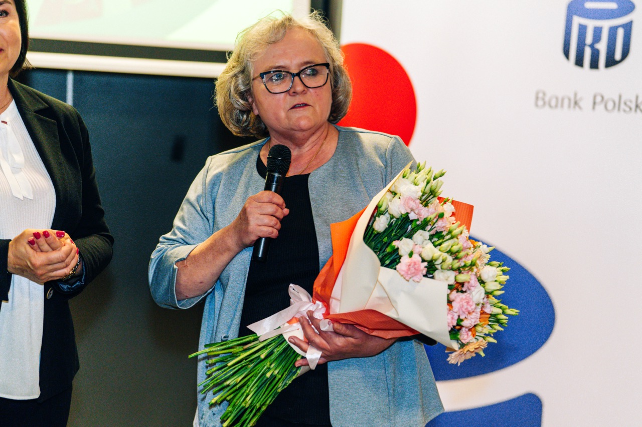 Uczestniczka rozdania wyróżnień PKO z bukietem kwiatów, mówiąca do mikrofonu w piwnicy hotelu.