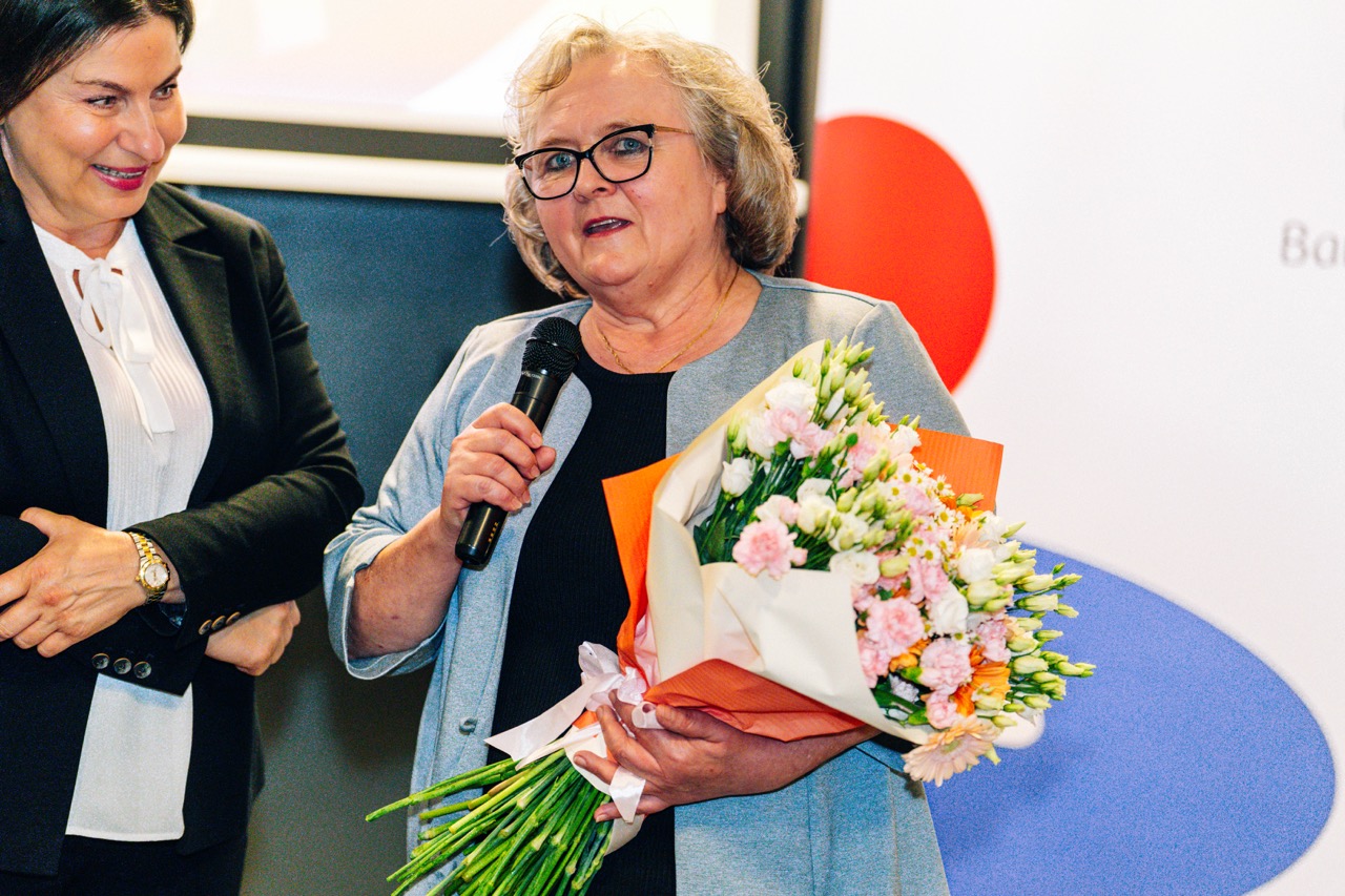 Uczestniczka rozdania wyróżnień PKO z bukietem kwiatów, przemawia w hotelowej piwnicy.