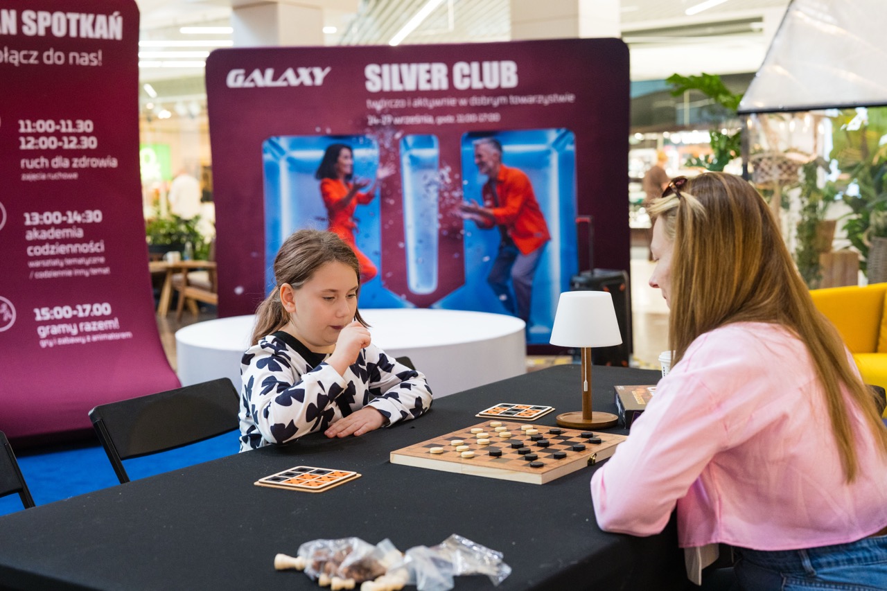 Uczestnicy warsztatów w CH Galaxy grają w warcaby, w tle baner Silver Club z informacjami o wydarzeniu.
