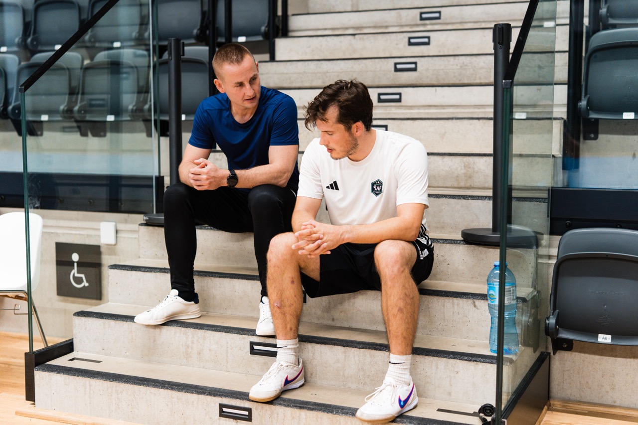 Trener i zawodnik rozmawiają siedząc na schodach hali sportowej