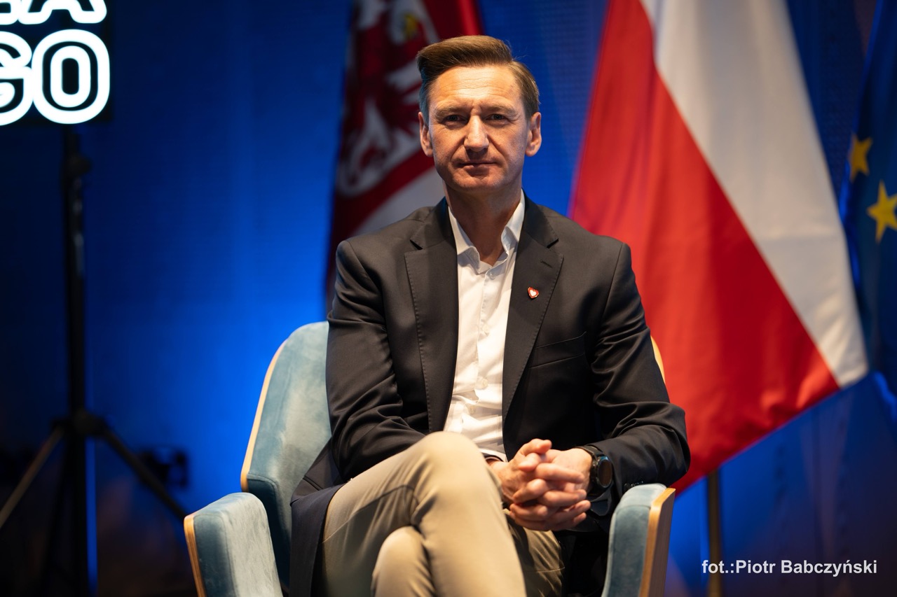 Prezentacja uczestnika wydarzenia BusinessContact6 w Szczecinie, siedzącego na scenie przed flagami.