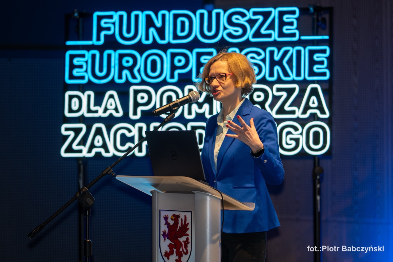 Prelegentka Alicja Defratyka podczas wystąpienia na konferencji BusinessContact6 w Szczecinie, z tłem neonowego napisu.