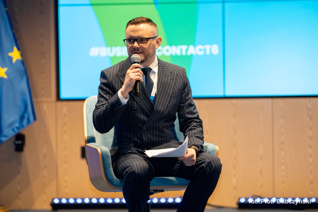 Prezentujący na scenie podczas BusinessContact6 w Szczecinie, trzymając mikrofon i notatki.