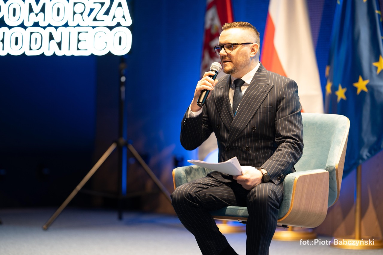 Prelegent w garniturze siedzi na krześle, prowadząc dyskusję podczas wydarzenia BusinessContact6 w Szczecinie.