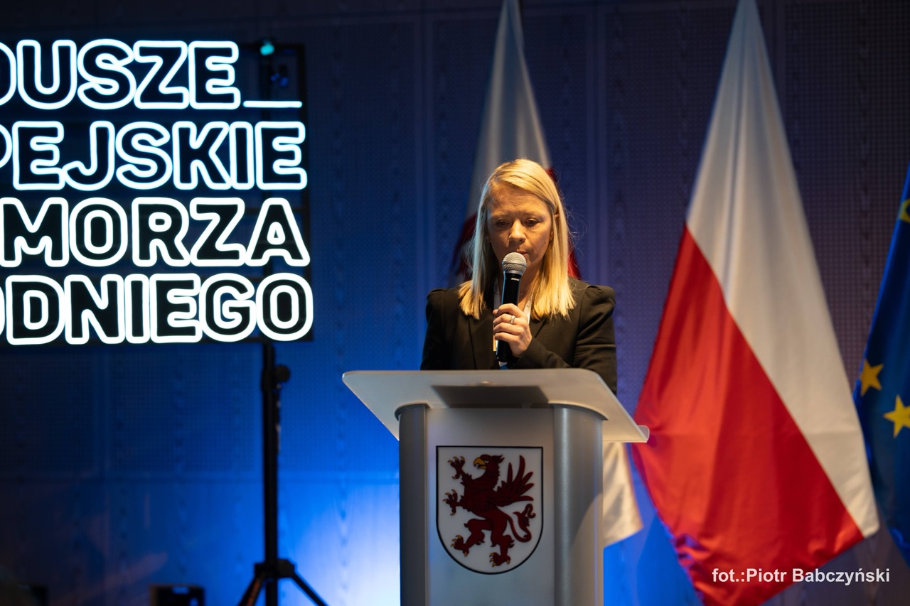 Fotografia eventowa z prelekcji w Szczecinie — Alicja Defratyka przy mównicy, z flagami w tle.