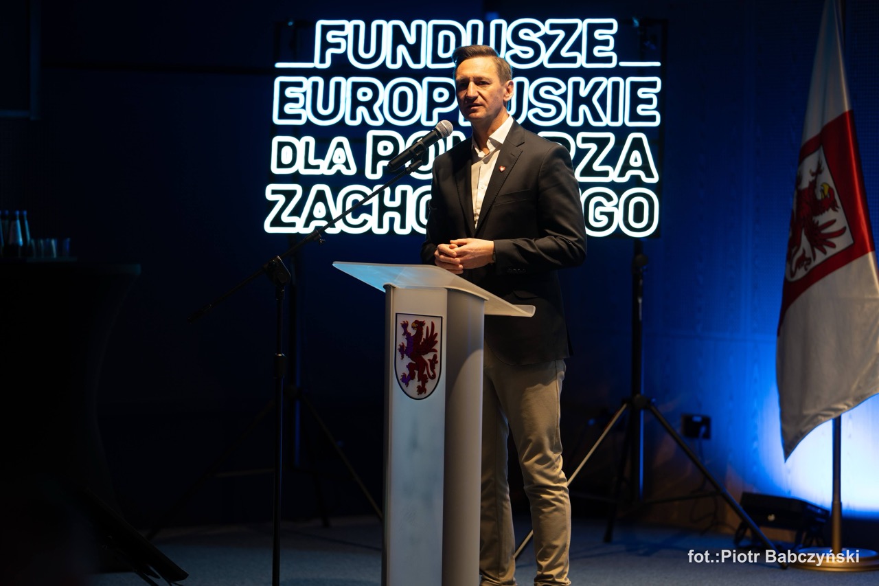 Prelegent na scenie podczas wydarzenia BusinessContact6 w Szczecinie, z neonowym napisem w tle.