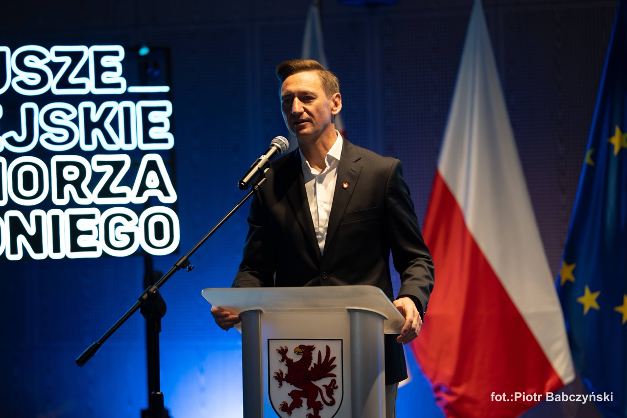 Prelegent przemawia z mównicy podczas konferencji BusinessContact6 w Szczecinie, z flagami w tle.