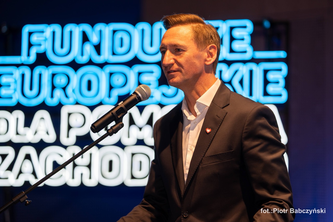 Prezentacja mężczyzny przy mikrofonie podczas wydarzenia BusinessContact6 w Szczecinie, z tłem w postaci podświetlonego napisu.