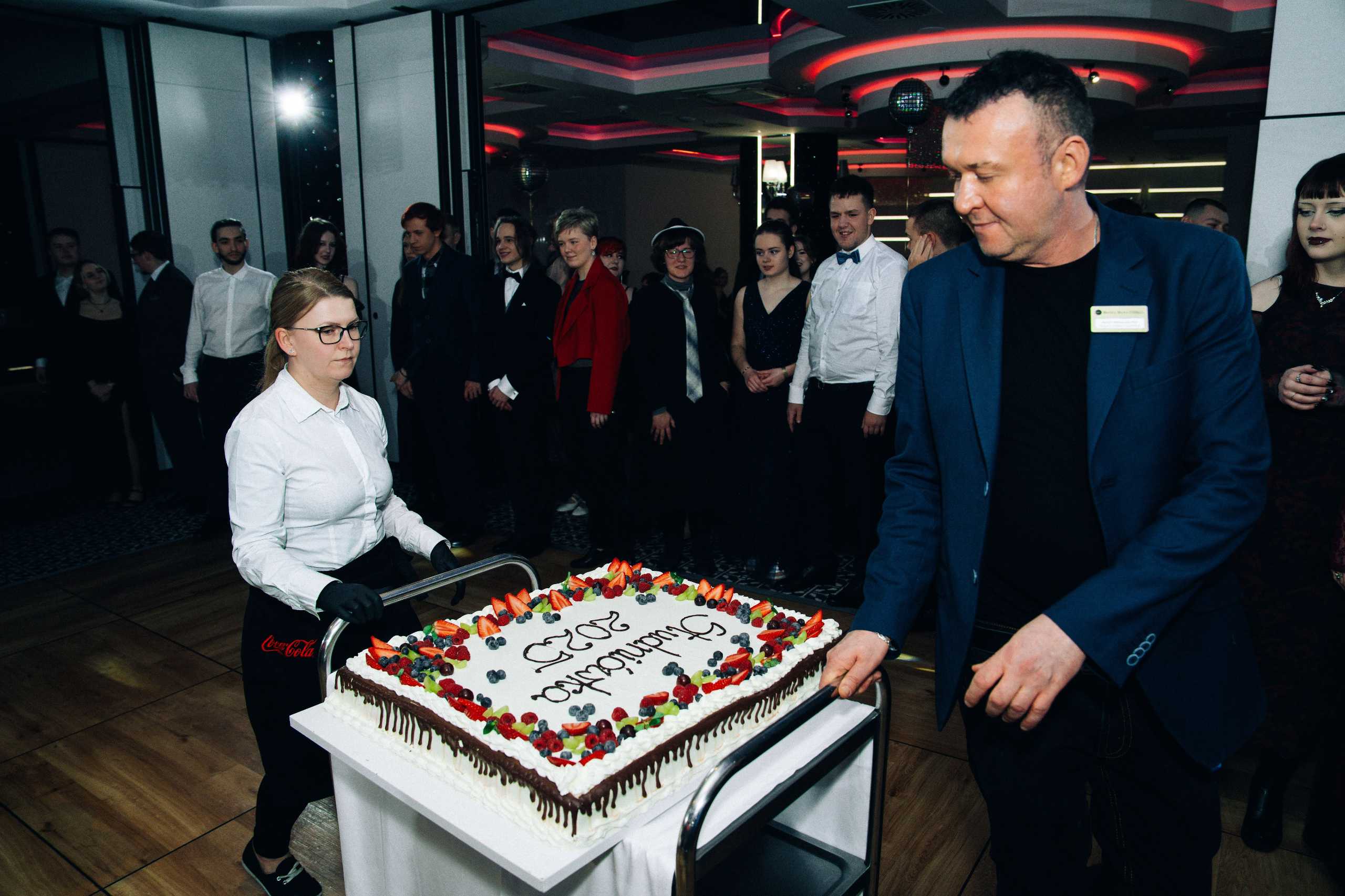 Moment serwowania tortu studniówkowego w Hotelu Dana, z uśmiechniętymi gośćmi w tle w Szczecinie.