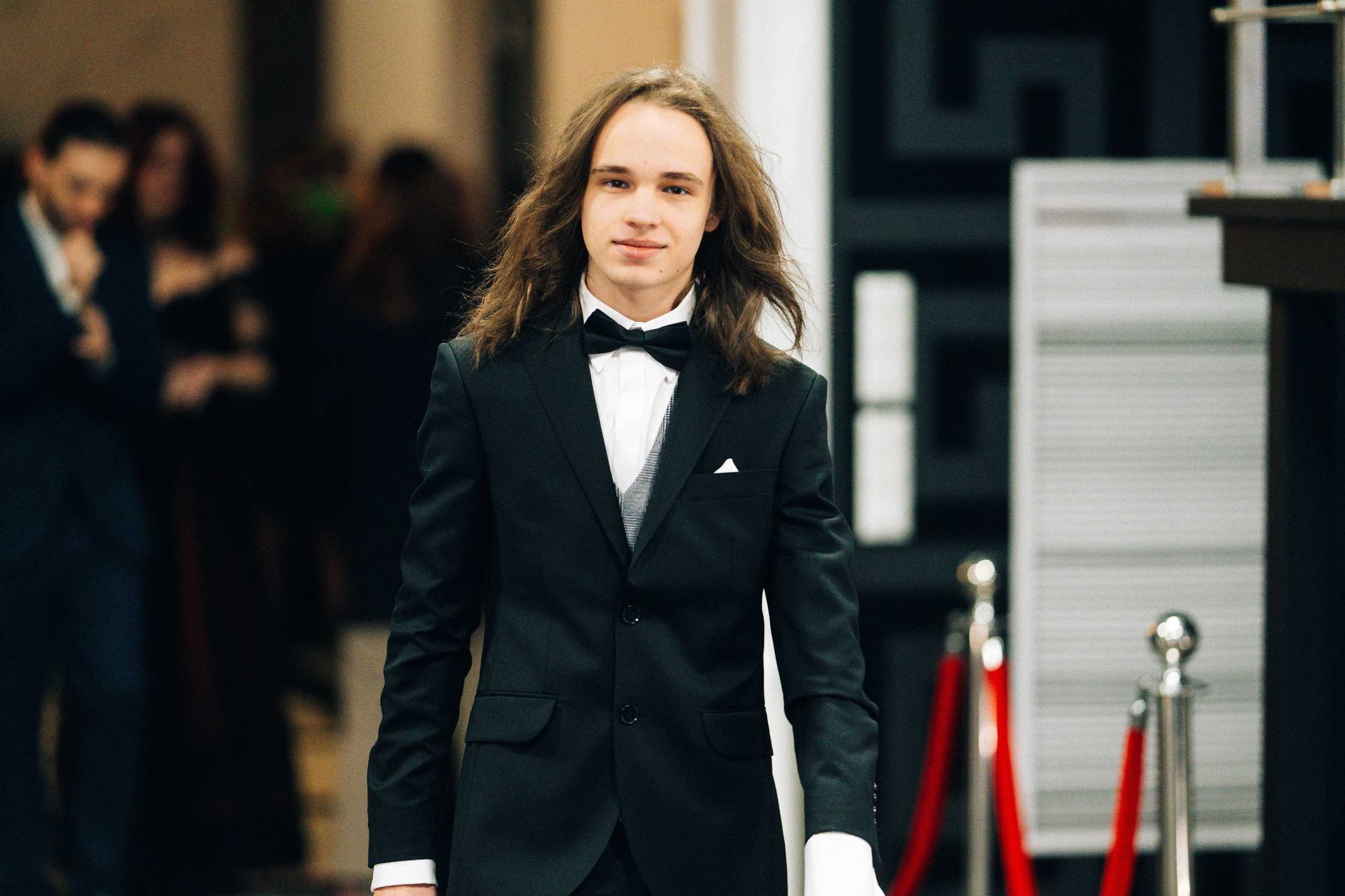 Młody mężczyzna w eleganckim garniturze z muszką, idący po czerwonym dywanie w Hotelu Dana w Szczecinie.